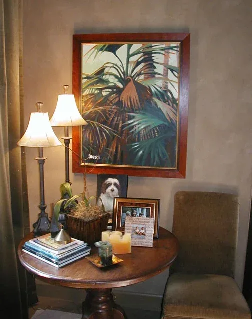 A cozy corner featuring a round wooden table with lamps, candles, books, and framed photos beneath a palm leaves painting.