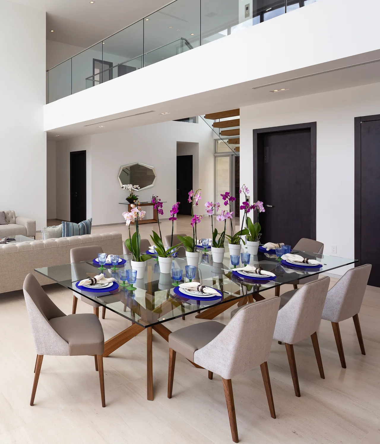 Dining area with glass table, upholstered chairs, and orchids under a balcony with glass railing.