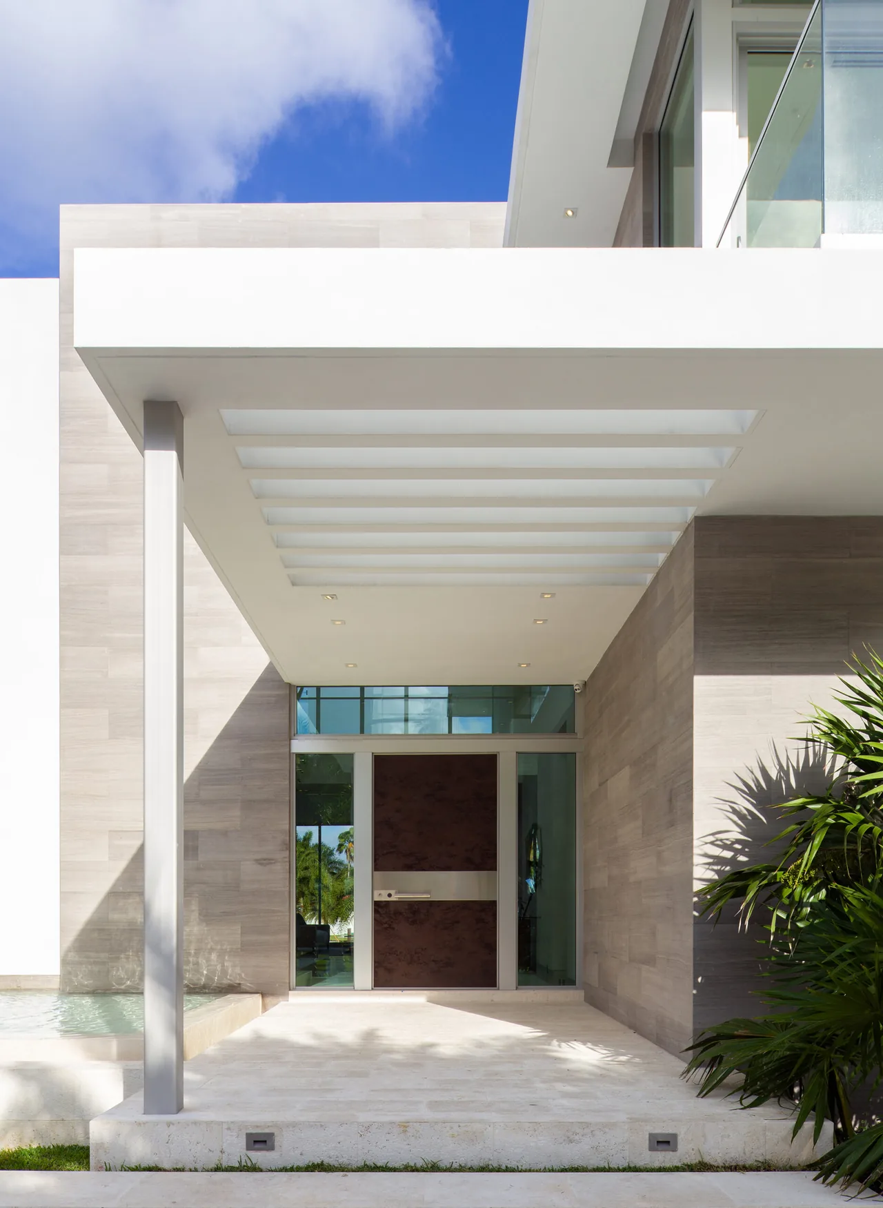 Entry of 301 Residence with a wide wood front door flanked by glass and a white overhang featuring skylights.