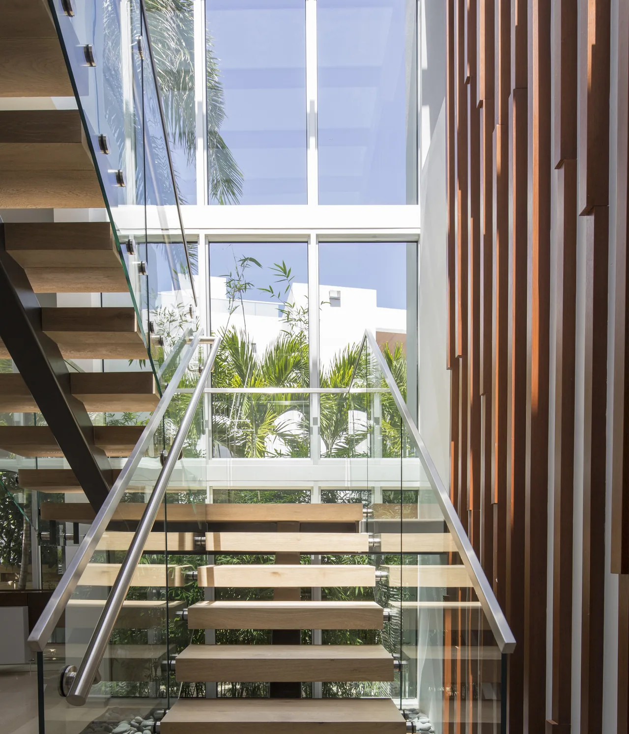 Floating wood stair treads supported by glass railing and metal handrails in a bright interior with vertical wood wall slats.