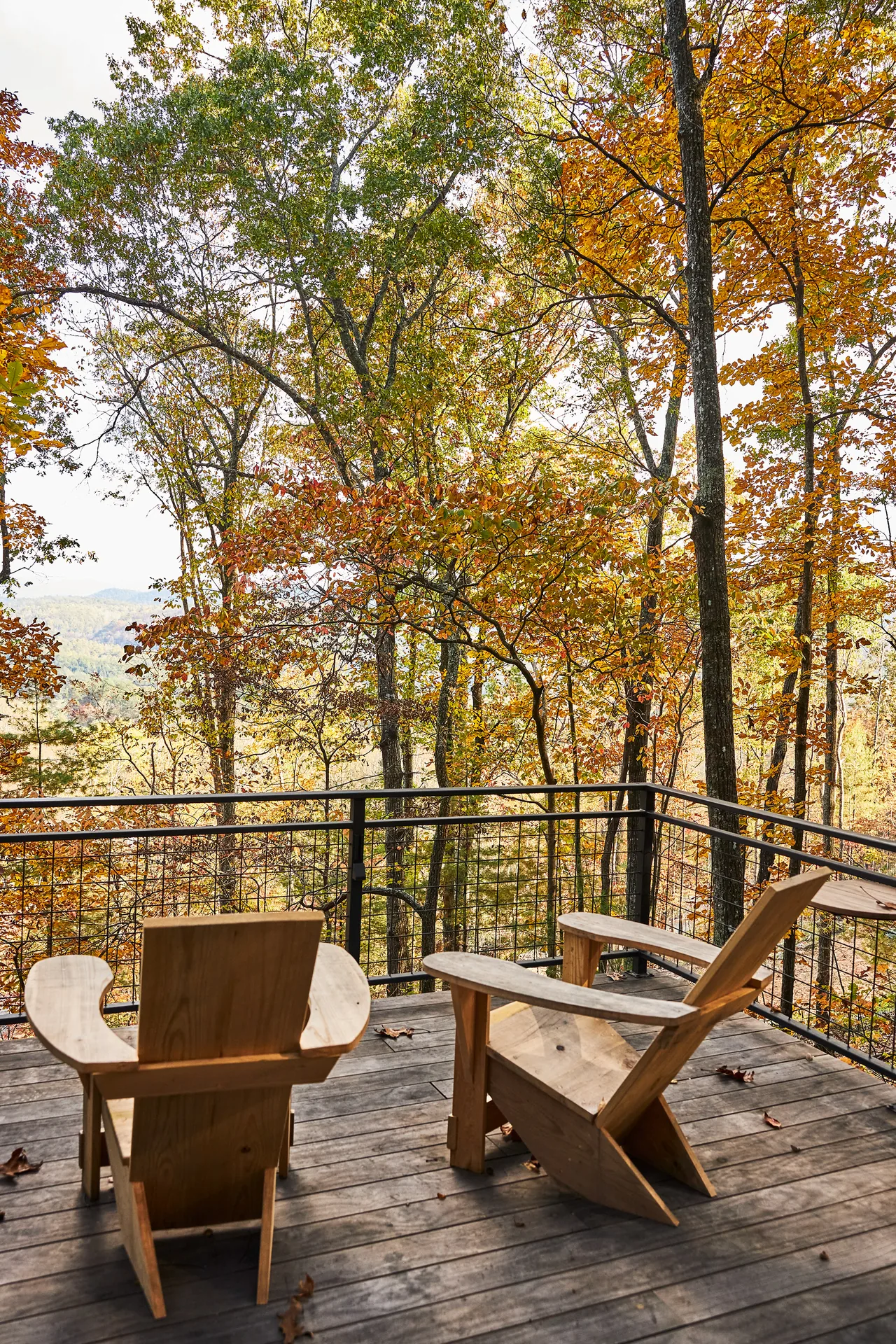 Deck with wooden chairs and metal railing overlooks colorful fall trees in a forest setting.