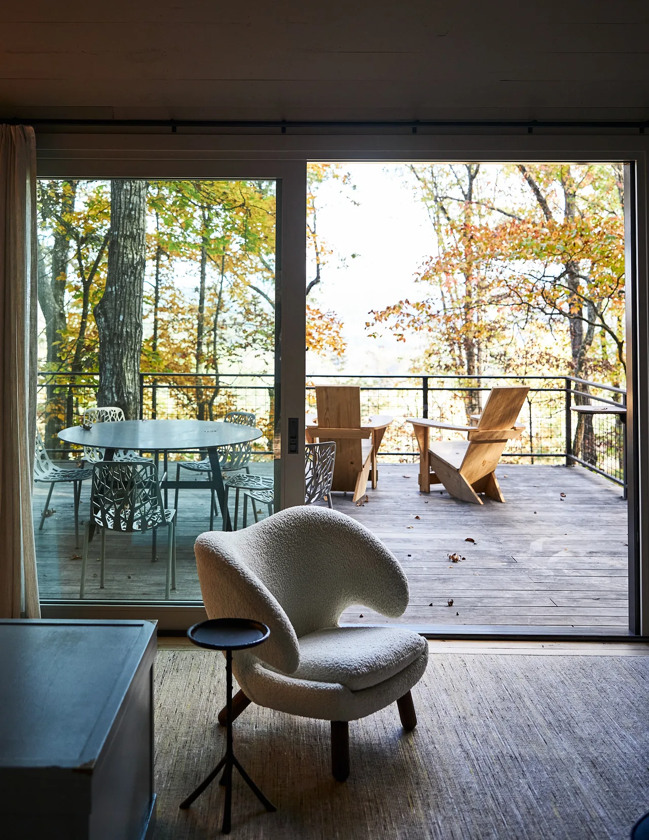 Living room with curved lounge chair and small table facing an outdoor deck surrounded by fall foliage