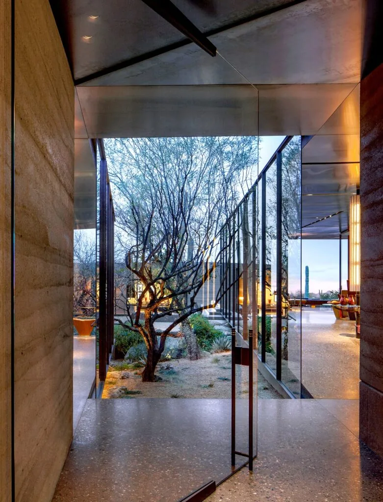 A rammed earth framed glass door opens to a desert courtyard with natural planting and integrated bench seating.
