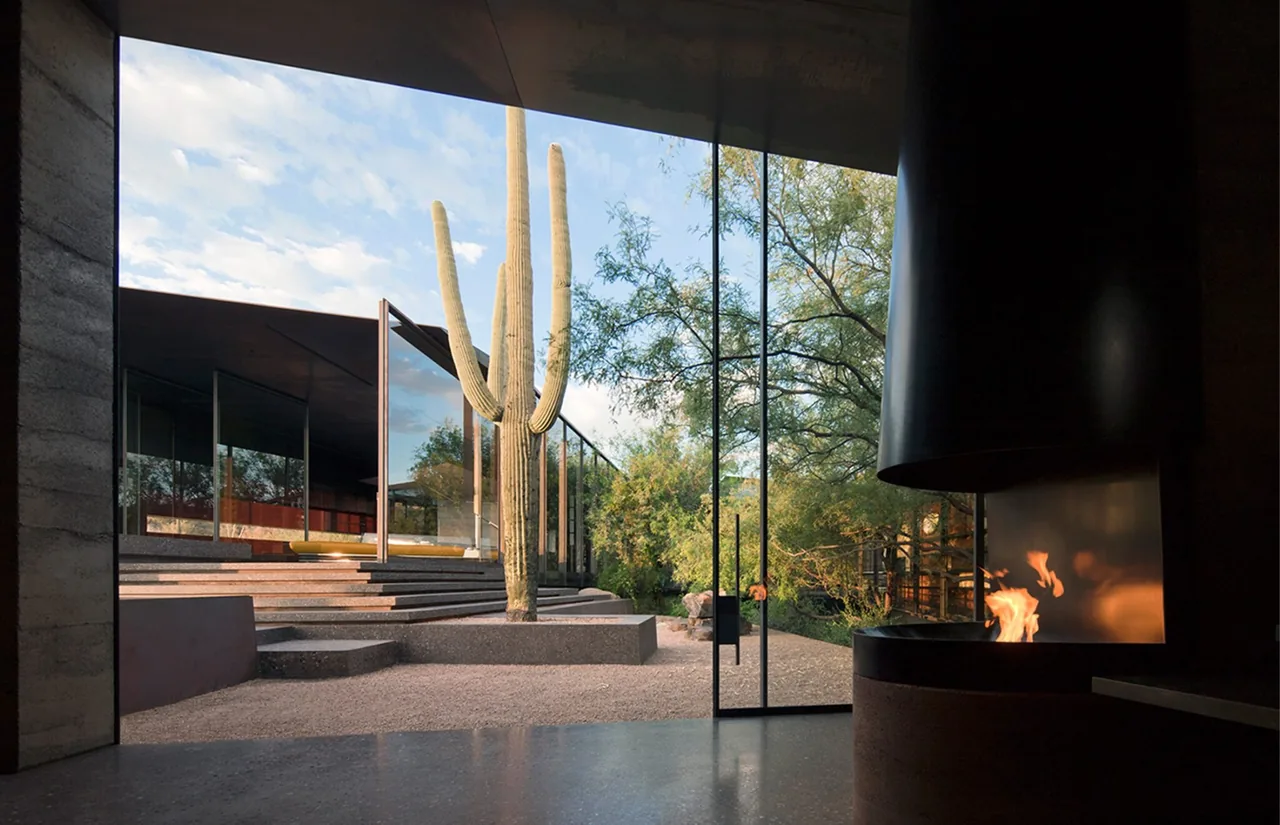 The Desert Courtyard House features floor-to-ceiling glazing overlooking a gravel courtyard with desert landscaping and an outdoor fireplace.