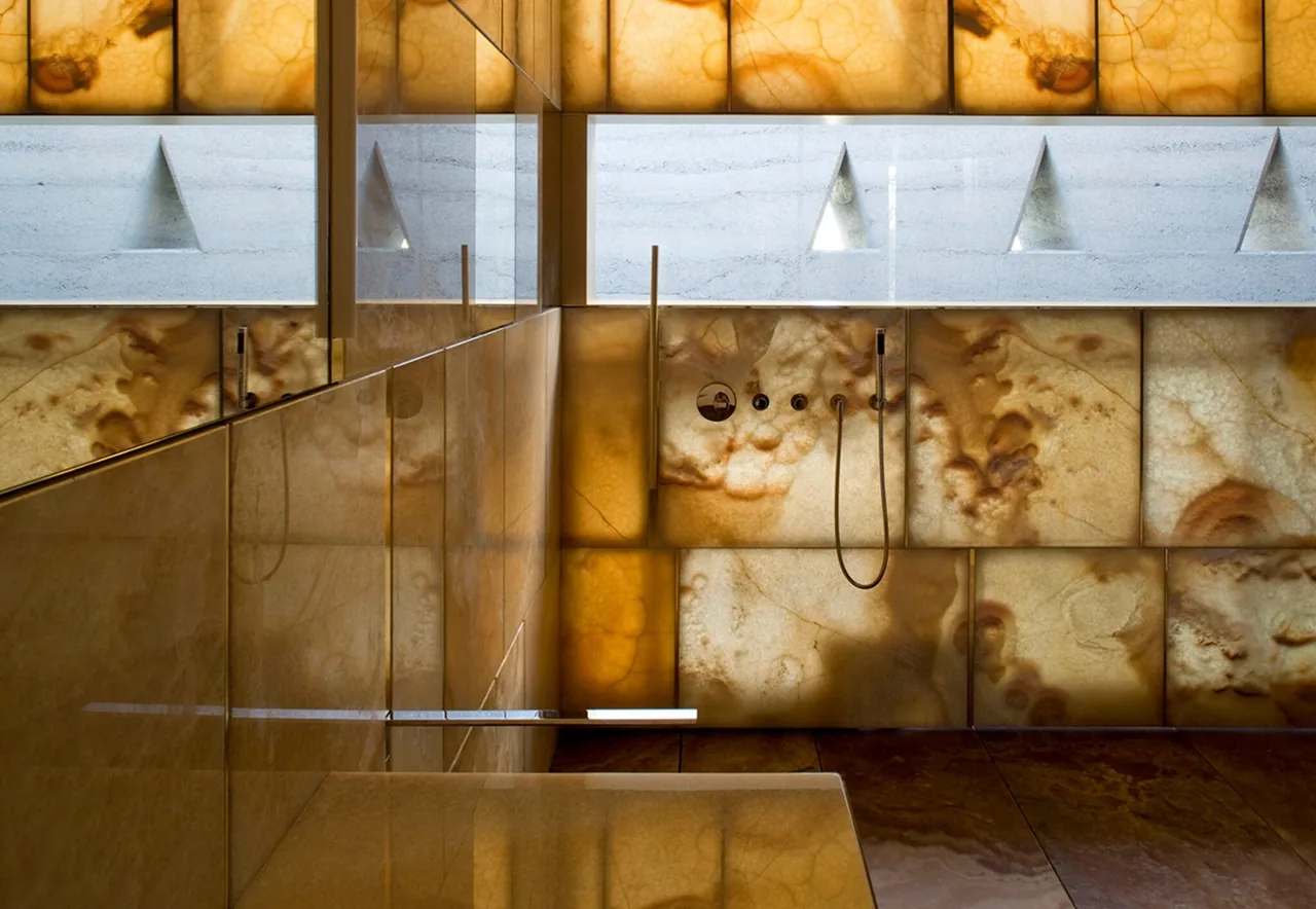 Shower area featuring backlit onyx or translucent stone tiles with a glass divider, and a narrow horizontal window above.