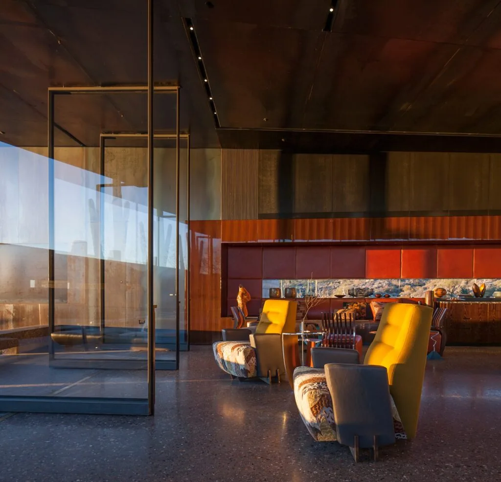 The Desert Courtyard House living area features large glass walls and a terrazzo floor with colorful chairs and custom wood details.