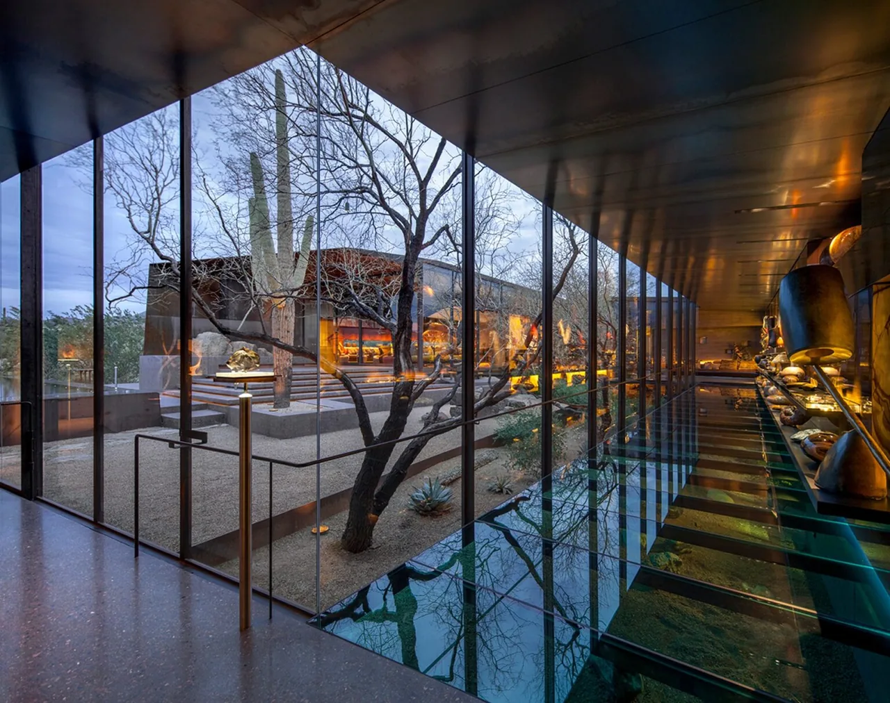 Interior corridor with floor-to-ceiling glass walls offering views of a desert courtyard with gravel landscaping and ambient lighting.