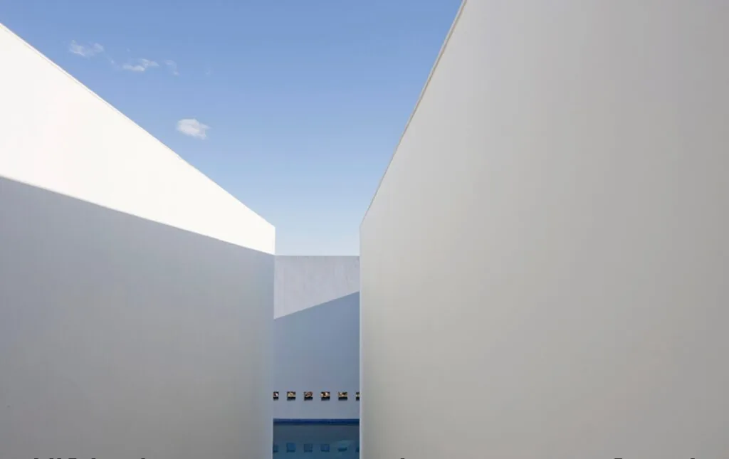 Minimalist exterior walls create a narrow passage leading to a pool area under a clear sky.