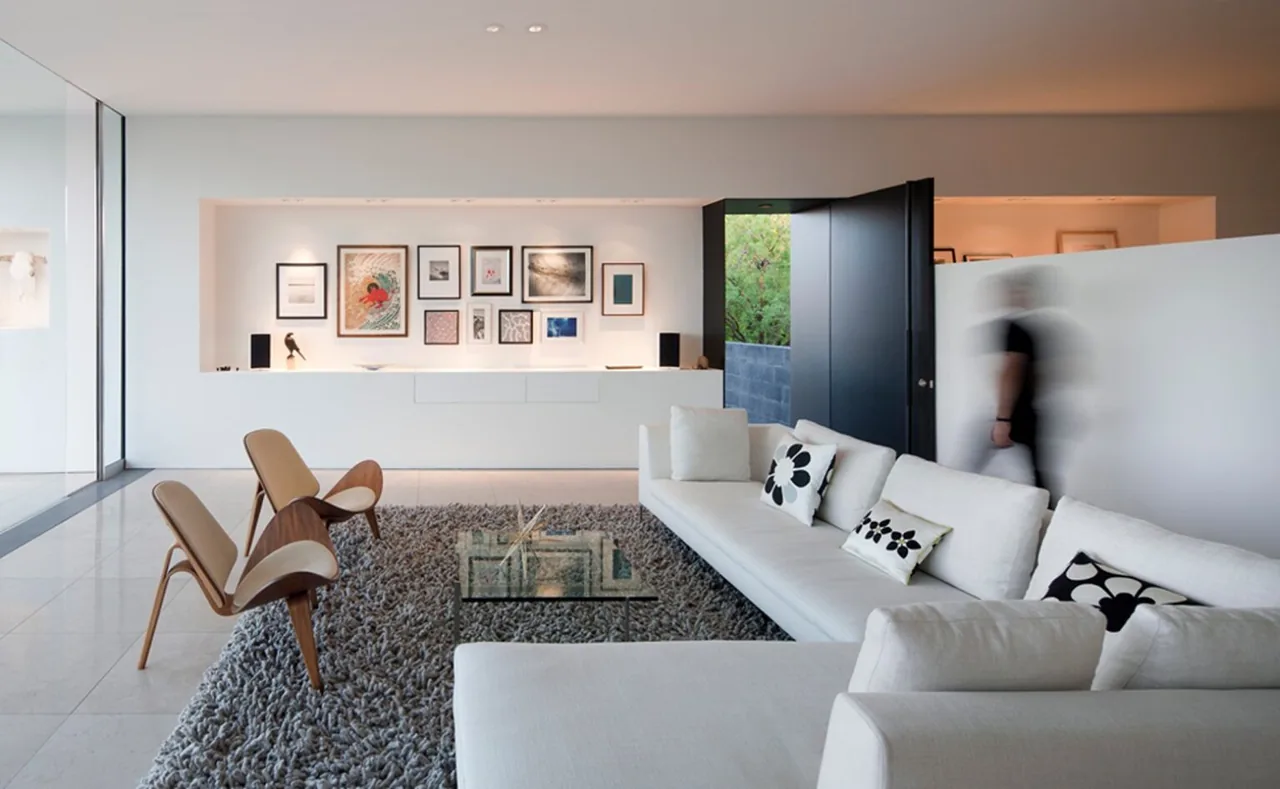 Dialogue House living room features a minimalist interior with a white sectional sofa, wood chairs, and framed artwork on a white wall.
