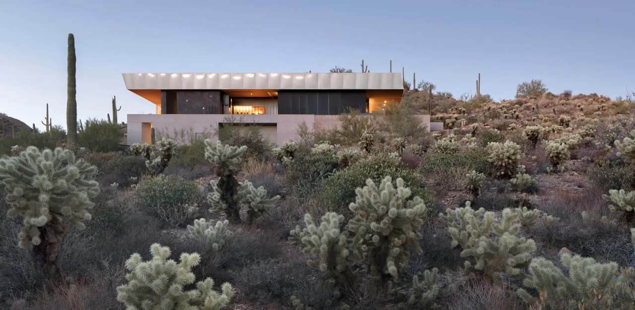 Modern flat-roof desert house with stacked stone walls situated on a hillside amid native desert plants.