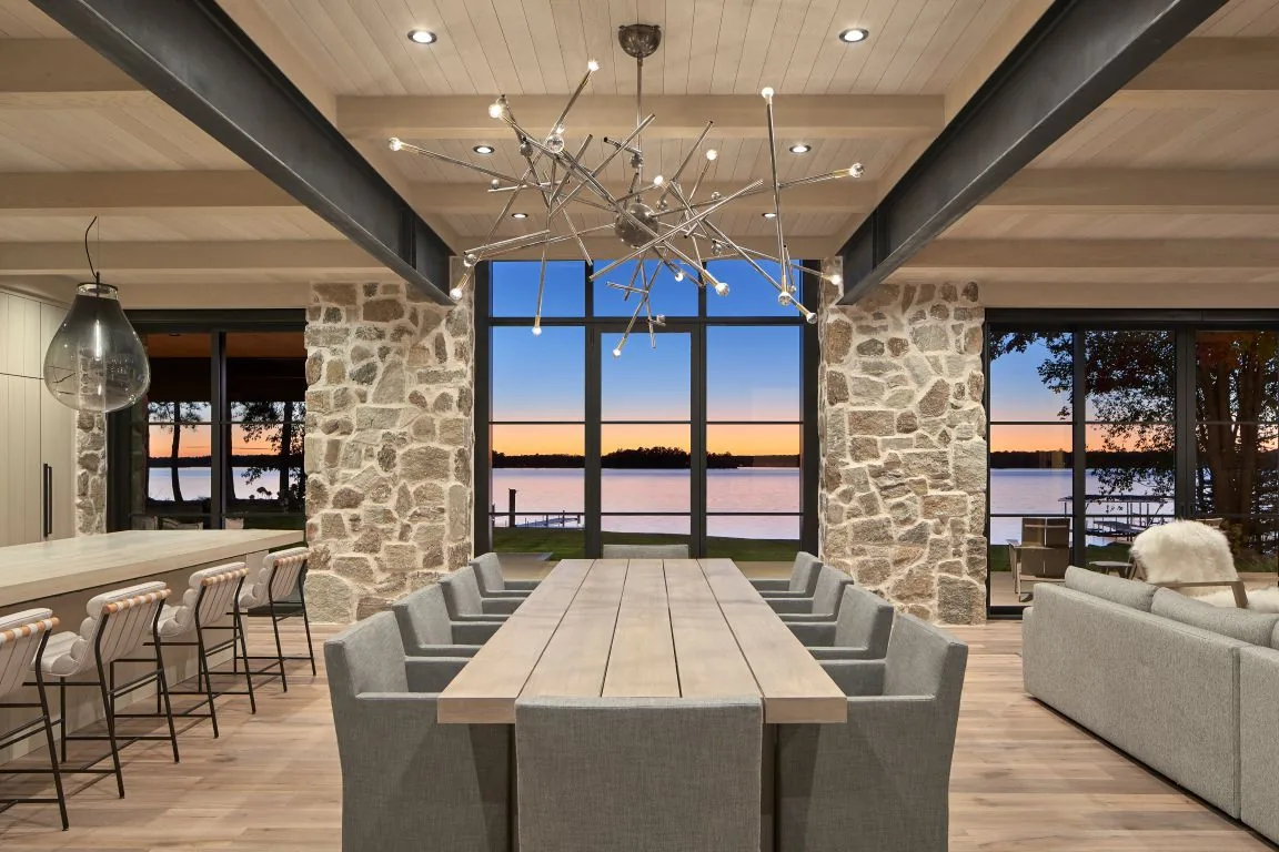 Dining area of Whitefish Lakehouse with stone walls, floor-to-ceiling windows, and a modern chandelier overlooking the lake at sunset.