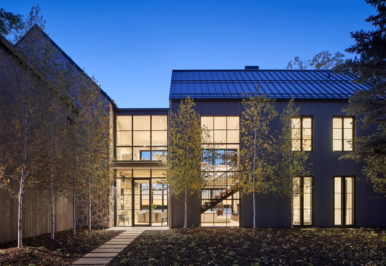 The Whitefish Lakehouse features large grid-style windows and a metal roof, illuminated at dusk to show interior elements.