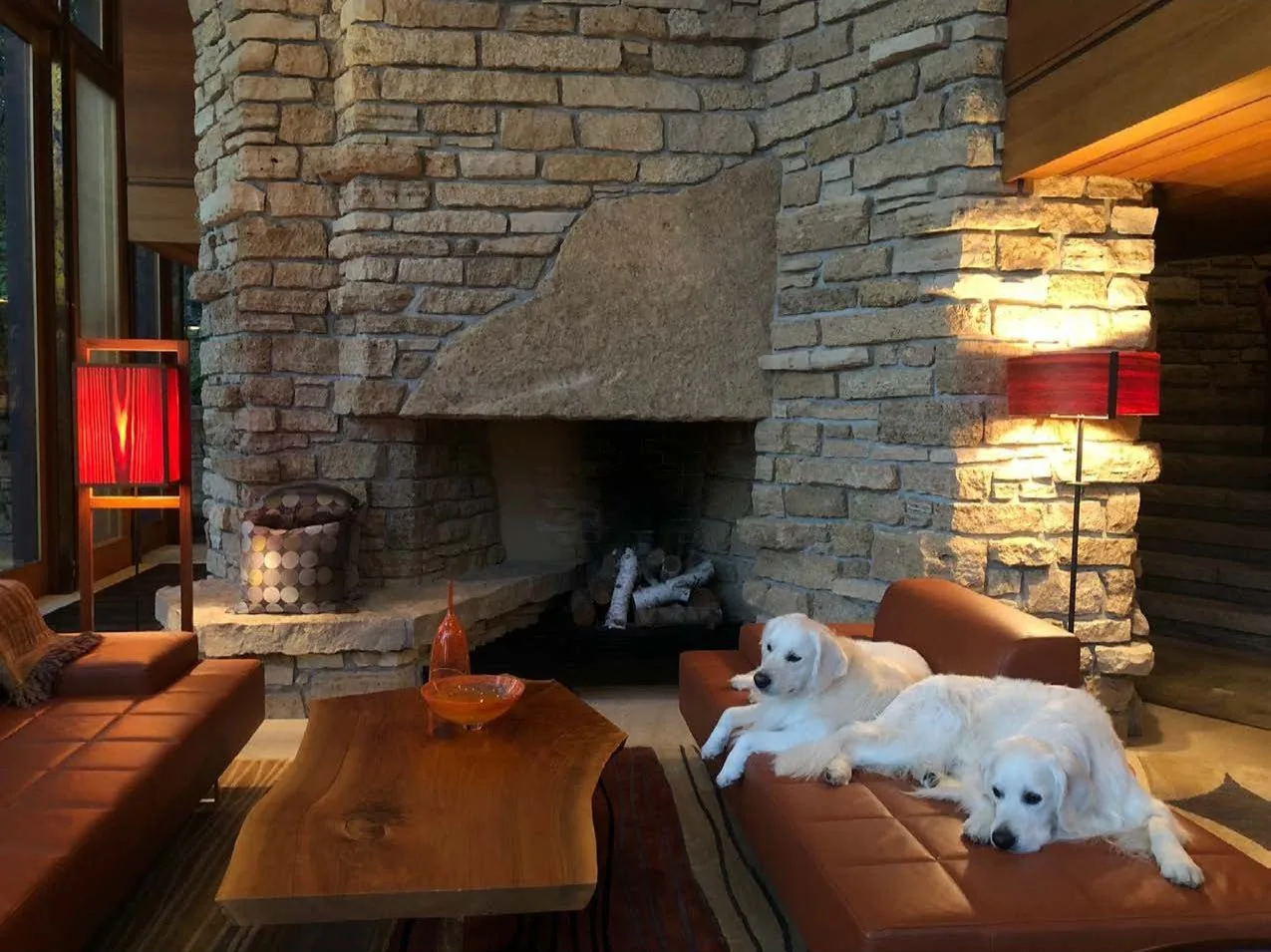 Living room features a large stone fireplace, brown leather sectional, unique wood coffee table, and warm ambient lighting.