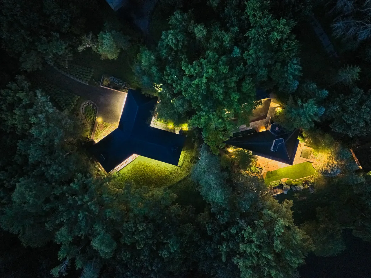 Night aerial photo of two houses with dark roofs nestled in a wooded setting and lit by exterior lights.
