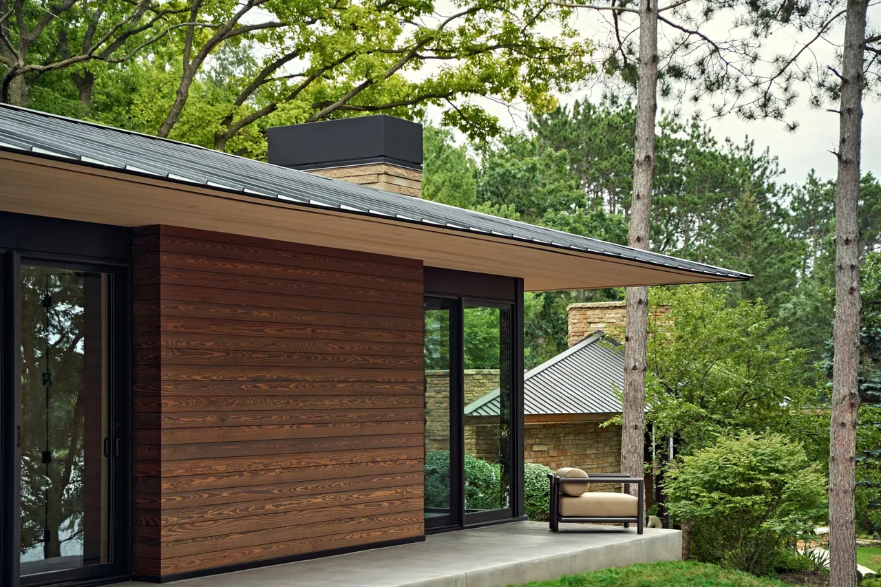 Modern home exterior with extended roof overhang, wood siding, black window frames, and sliding glass doors adjacent to a forest setting