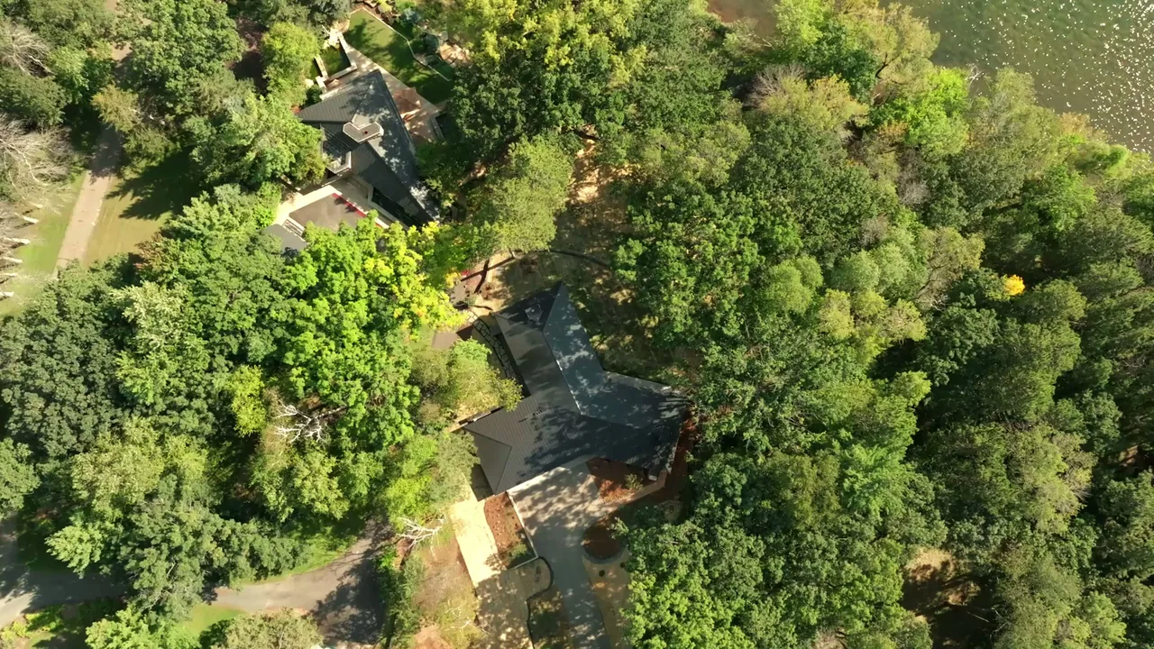 Aerial image shows two houses with dark roofs nestled in a lush forest setting near a pond or lake.