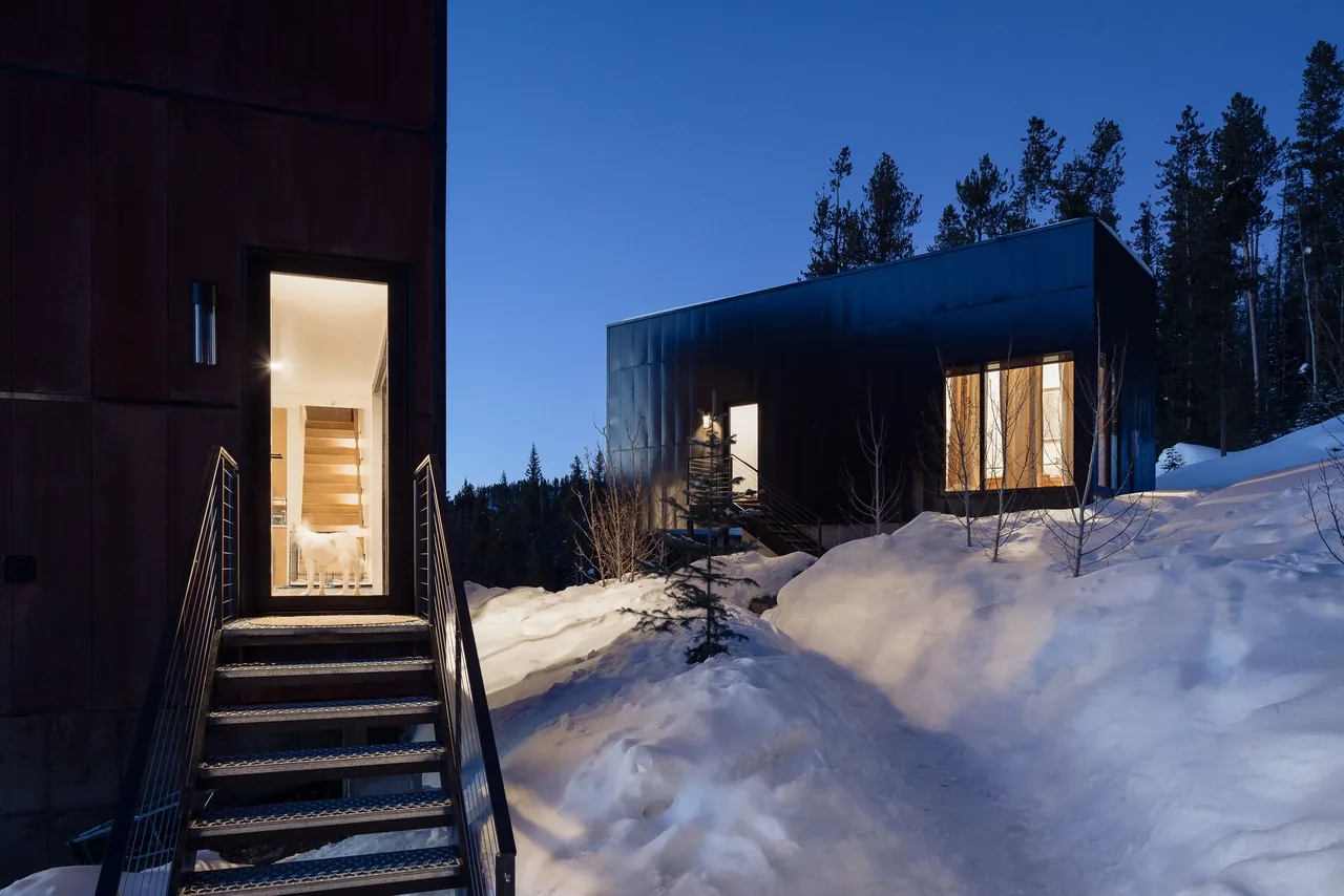 Two modern metal-clad buildings stand on a snowy landscape at dusk, lit warmly from within.