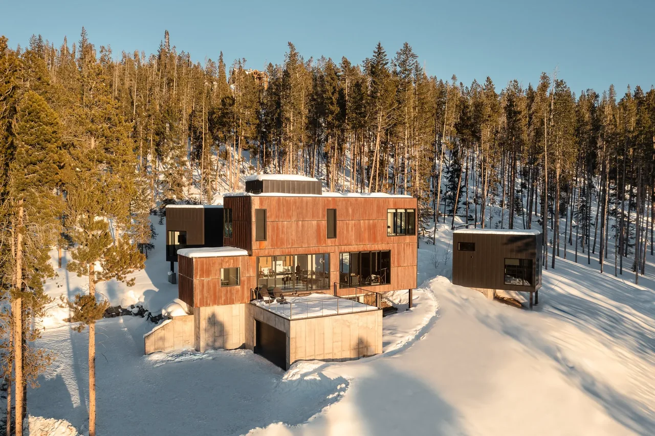 Modern two-part home with rust and dark siding on a snowy mountain slope amid tall pine trees.