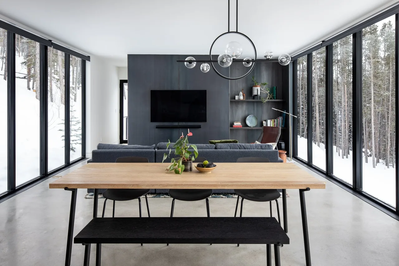 Modern open living and dining space with large black window frames and a view of snow-covered trees.