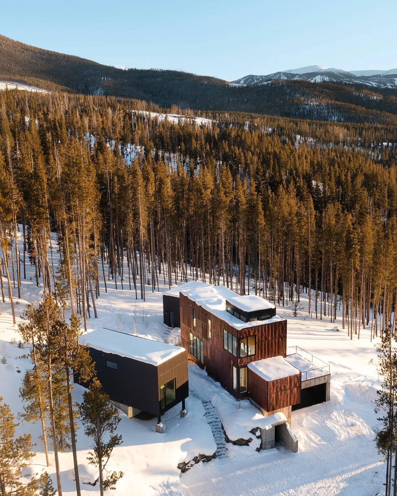 Modern two-part home in a snowy forested mountain setting with exterior stairs connecting the structures.