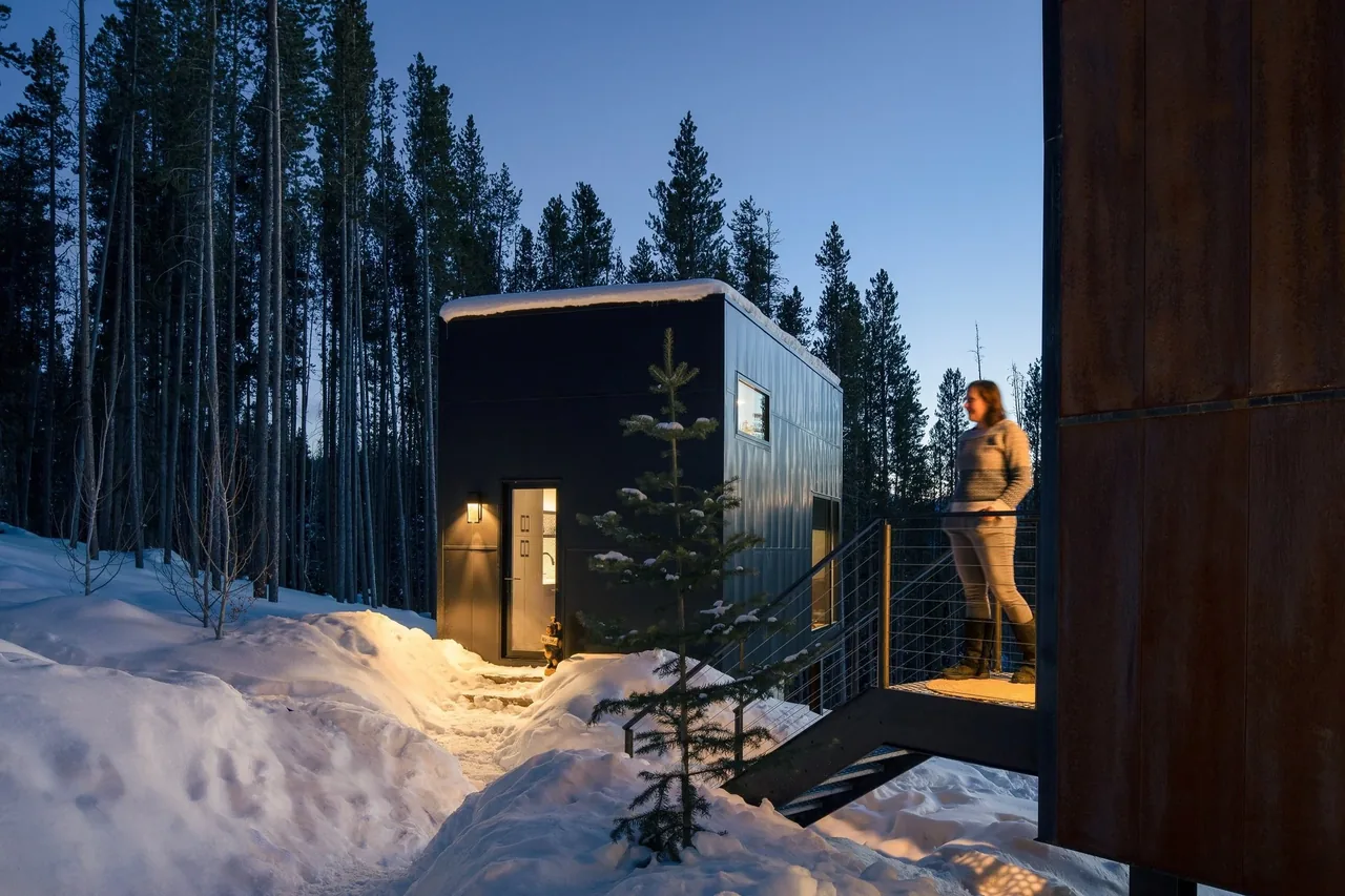 Small modern black metal siding building in a snowy forest at dusk with exterior lighting and adjacent elevated walkway.
