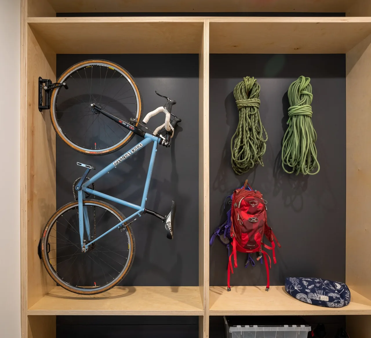 Wooden cubby storage displays a vertically mounted blue bicycle, climbing ropes, and a red backpack.