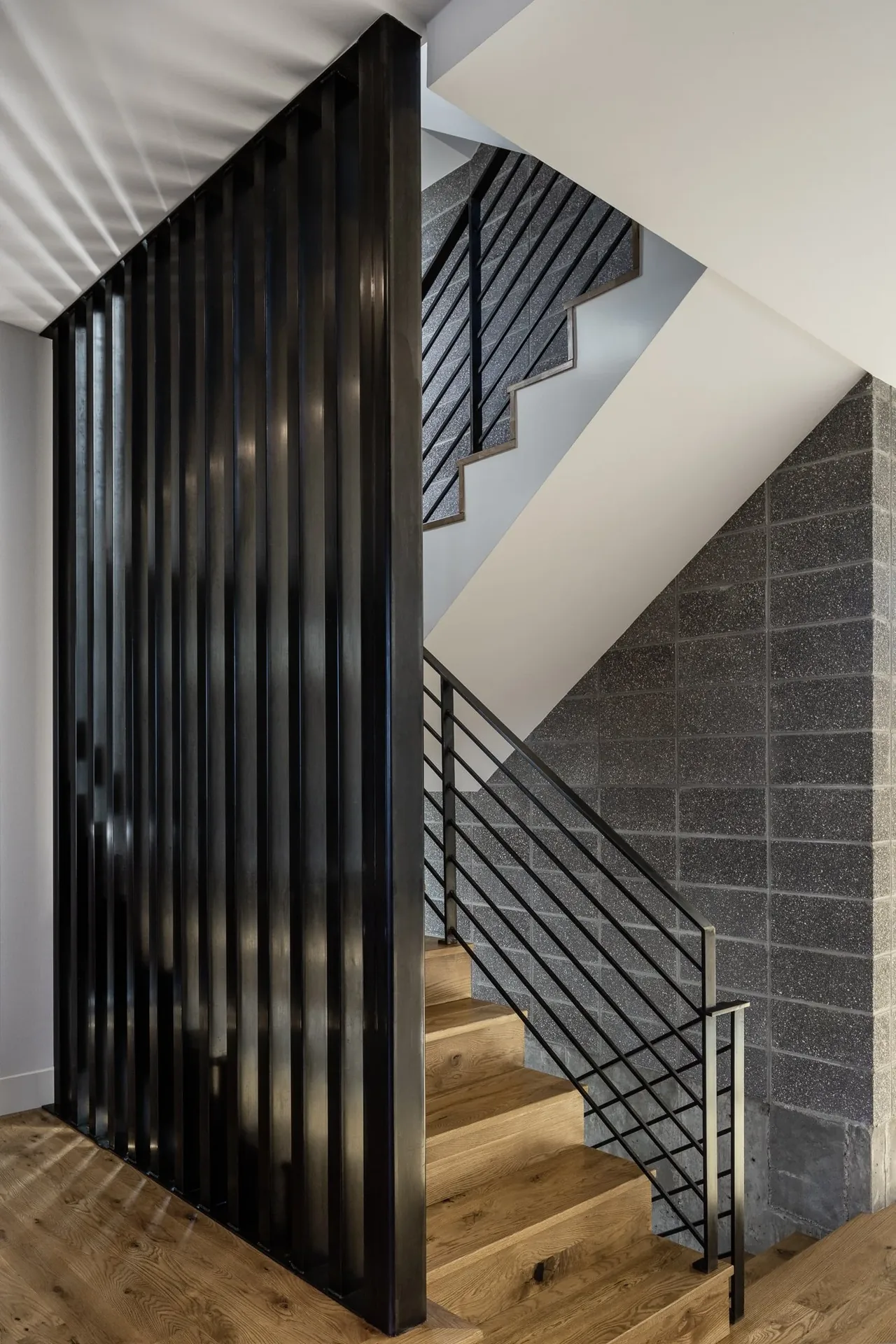 Interior wooden staircase with black metal railing and vertical slat divider beside a gray concrete block wall.
