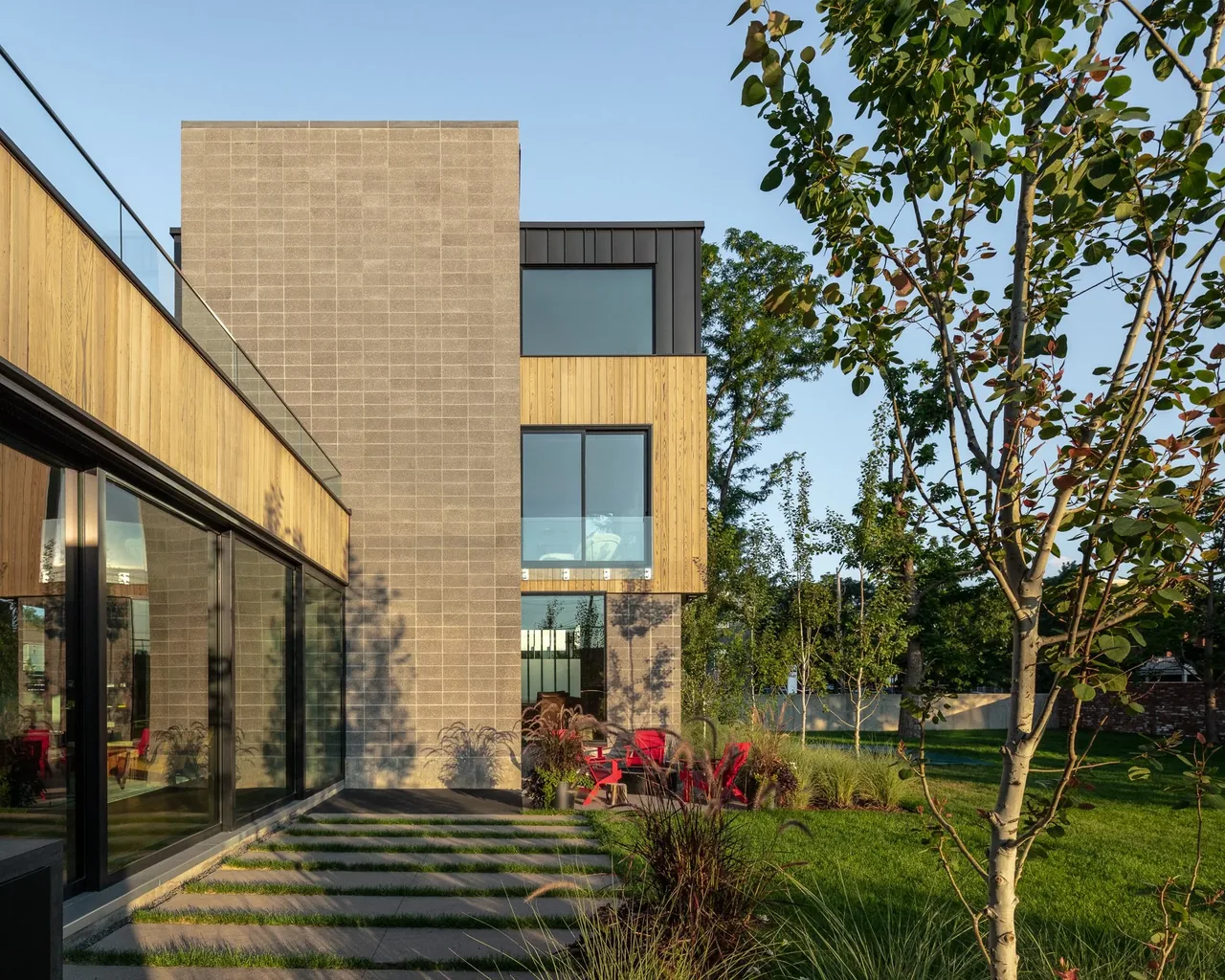 Modern residence exterior features wood and brick siding with large windows and a landscaped grassy yard including red outdoor seating.