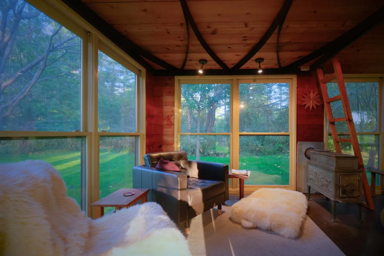Small playhouse interior with cast iron stove, ladder access, and large windows providing natural light and views of the outdoor greenery.