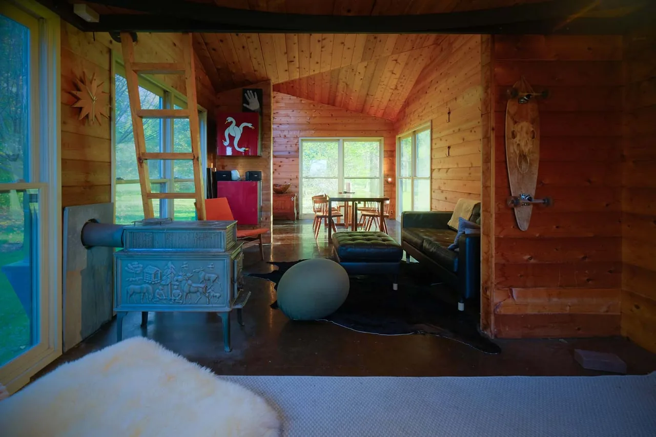 Cozy wood-paneled interior of a small playhouse featuring a cast iron stove and lounge furniture with natural light from multiple windows.