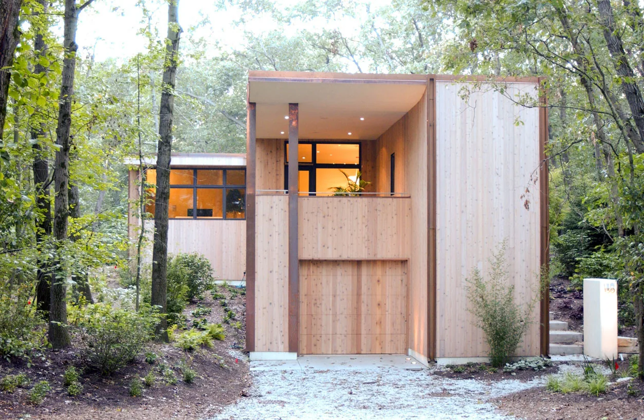 The McAllister Road House features wood siding, large black window frames, and a covered entry balcony in a forested setting.