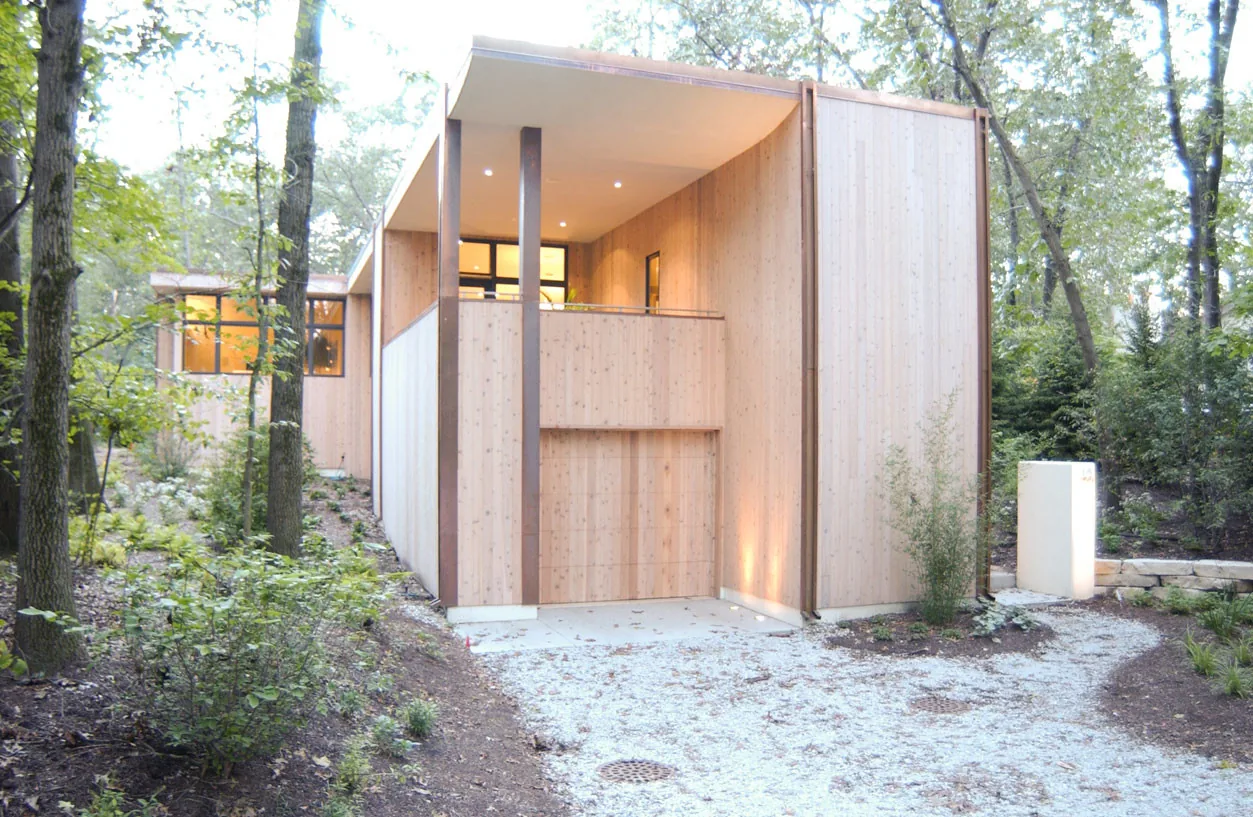 The McAllister Road House features vertical wood siding and a covered entry balcony in a wooded environment.