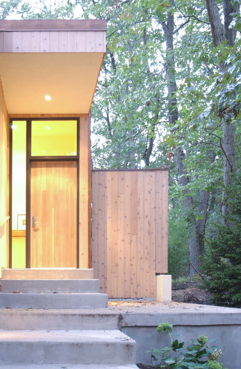 Modern front entry of the McAllister Road House with vertical wood siding and concrete steps in a forest setting.