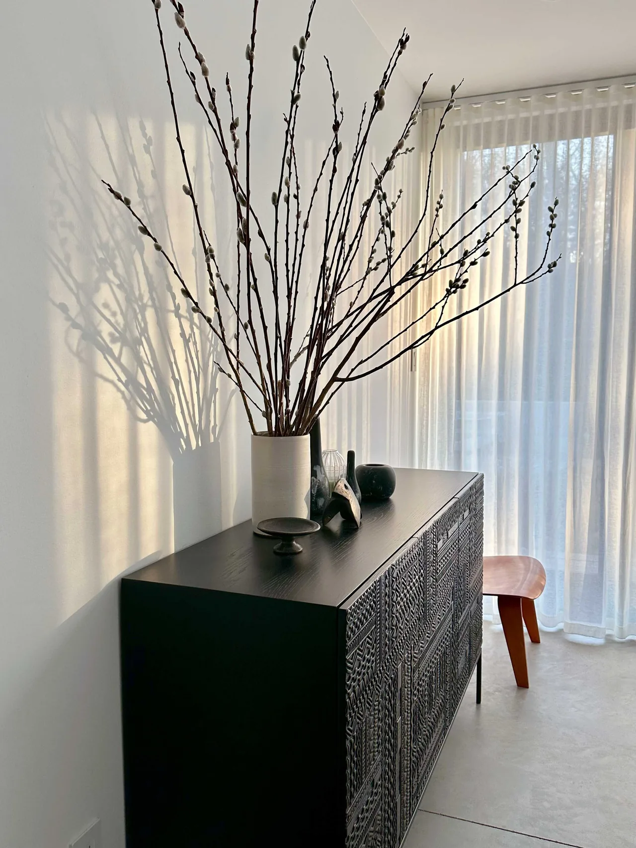 Carved black cabinet with decorative objects and tall branches next to large sheer curtained windows.