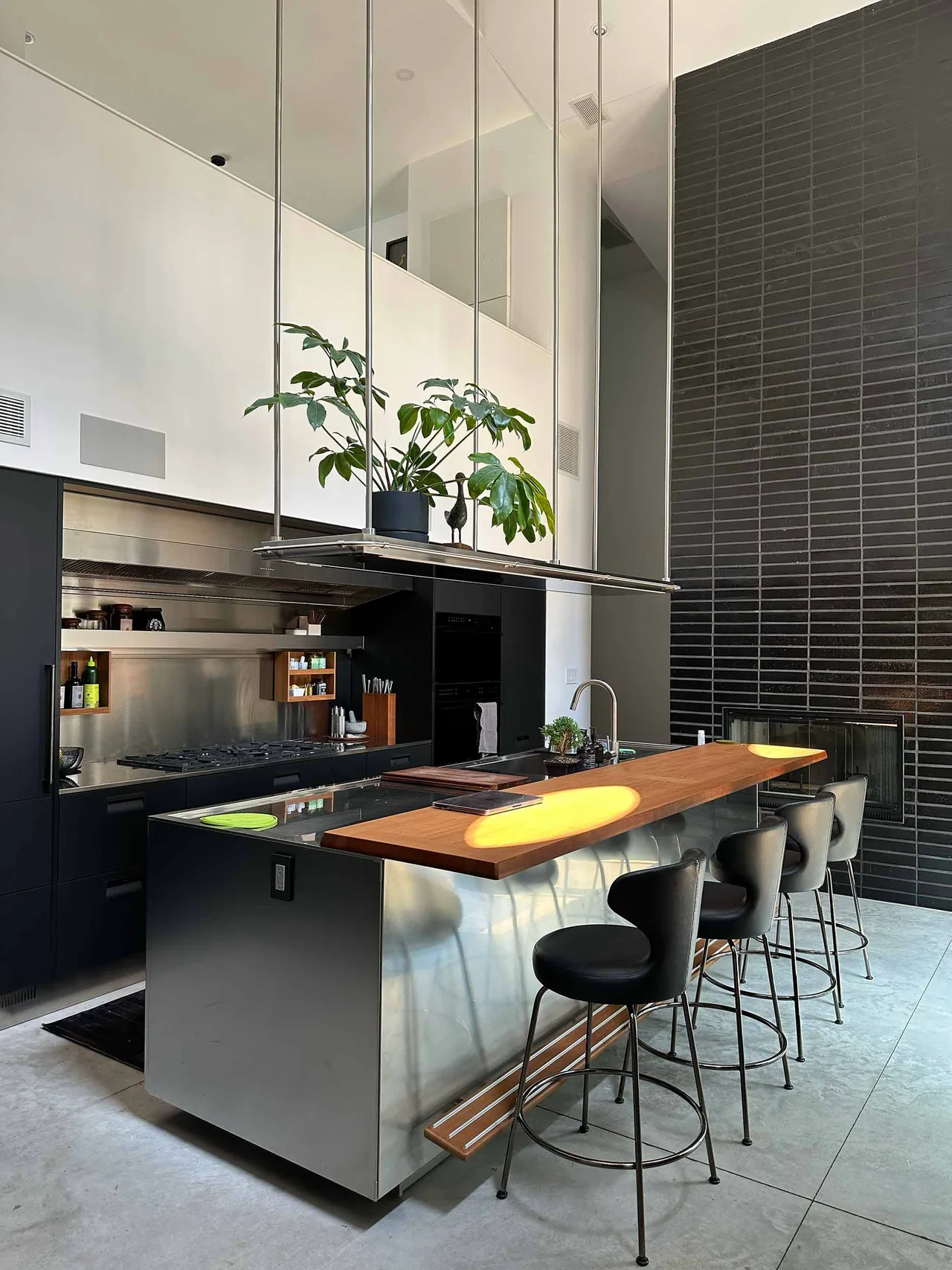 Contemporary kitchen features a stainless steel island with wooden bar counter and black stools, set against a tall black tiled fireplace wall.