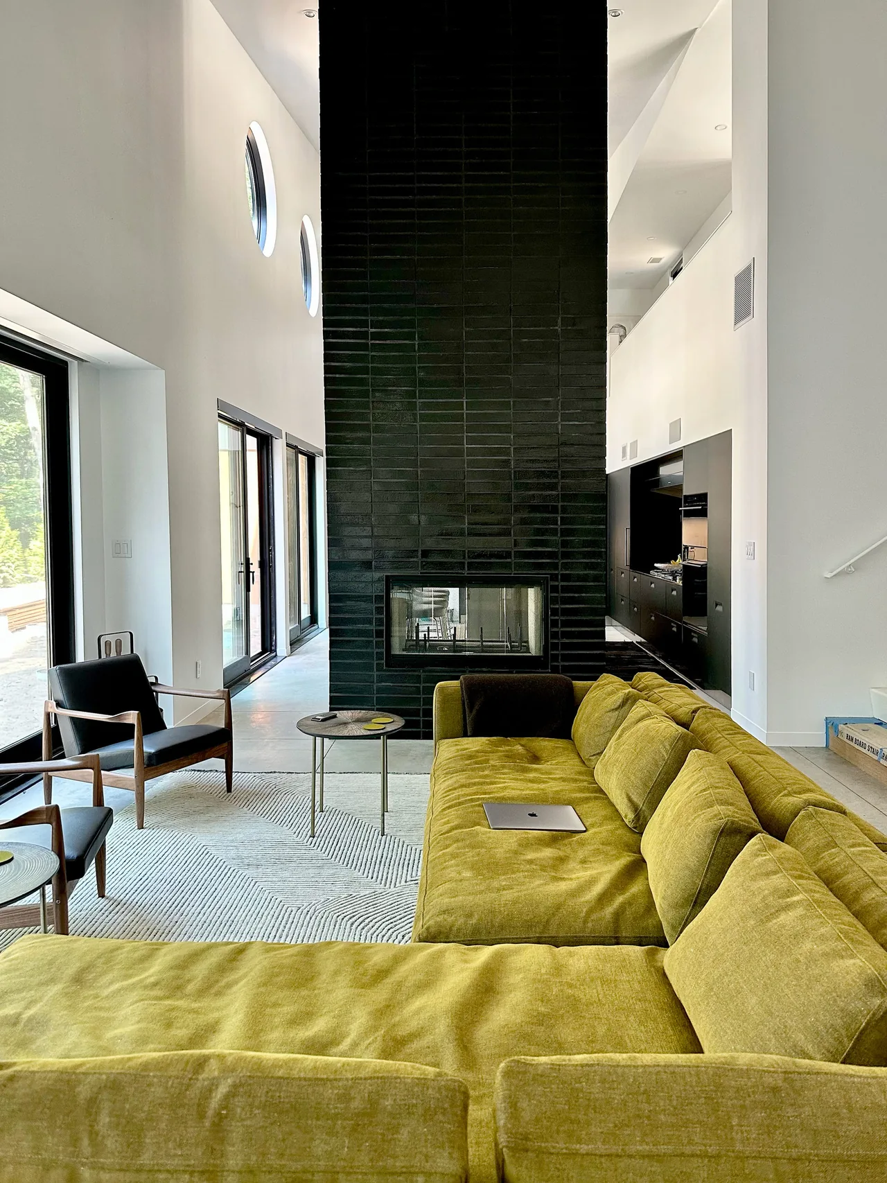 Modern living room with a large olive green sectional and a tall black tile double-sided fireplace dividing the space from the kitchen.