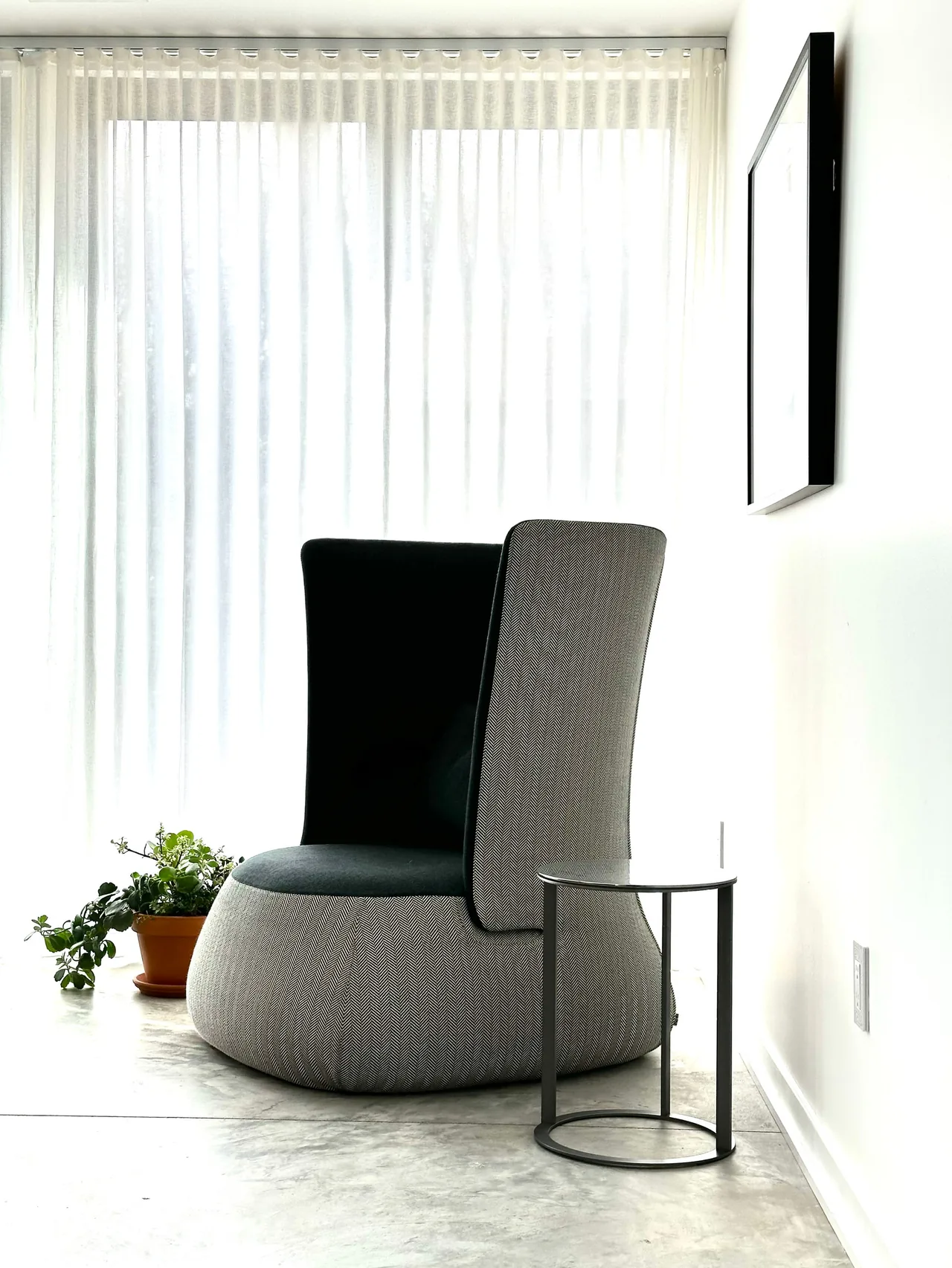 This cozy lounge chair with a rounded base and small side table is placed near sheer curtains in a bright room.