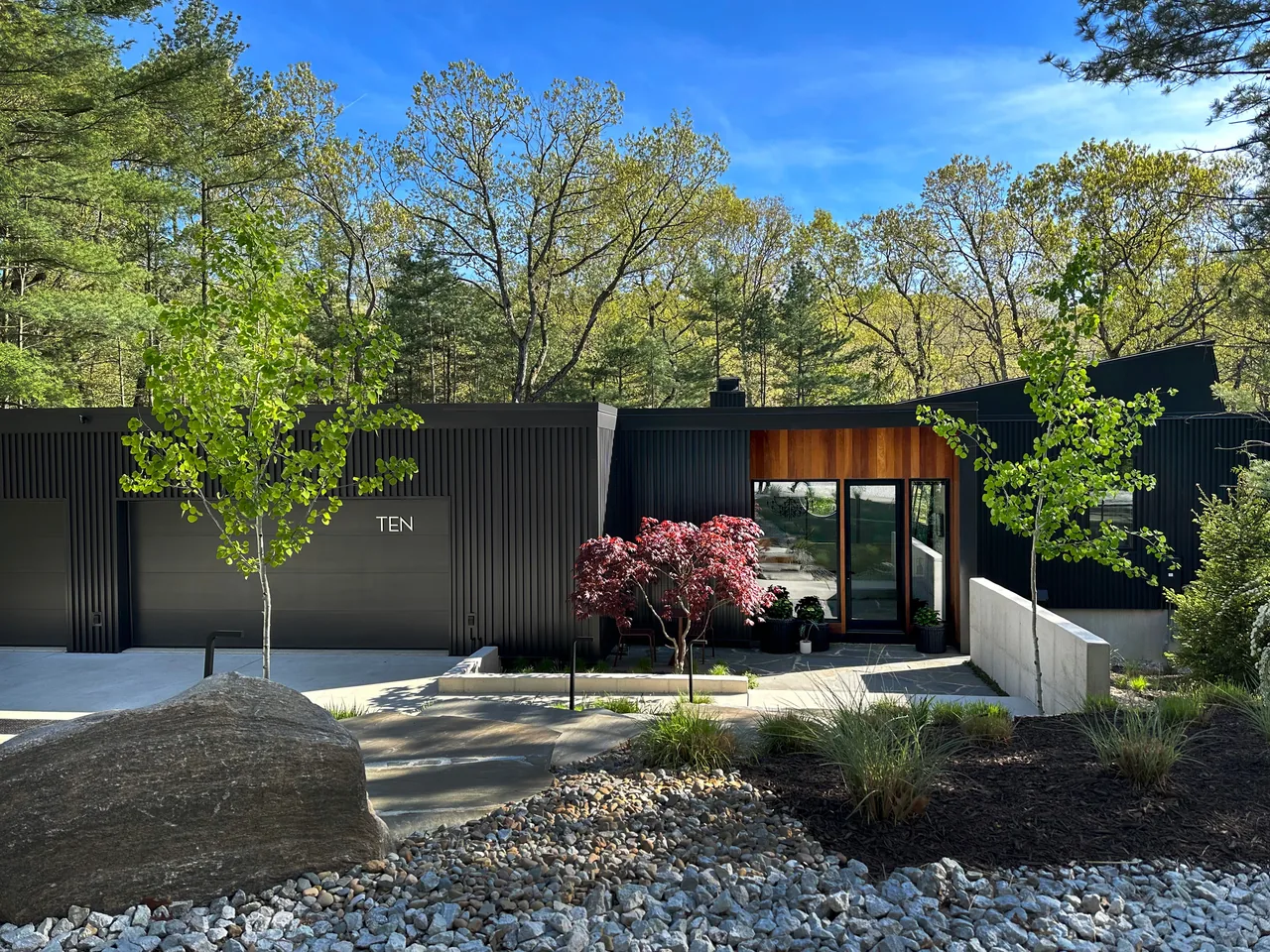 Modern Dune features black metal siding, wood-clad entry, and stone steps amid a natural landscape with ornamental grasses.