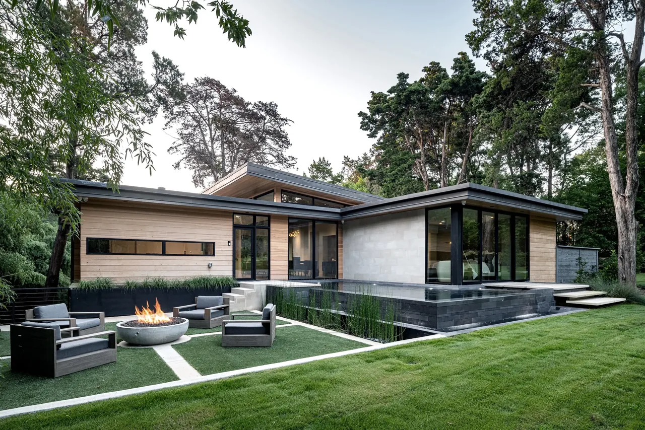 Modern bungalow exterior rear with flat roof, black window frames, firepit seating, and integrated reflecting pool.