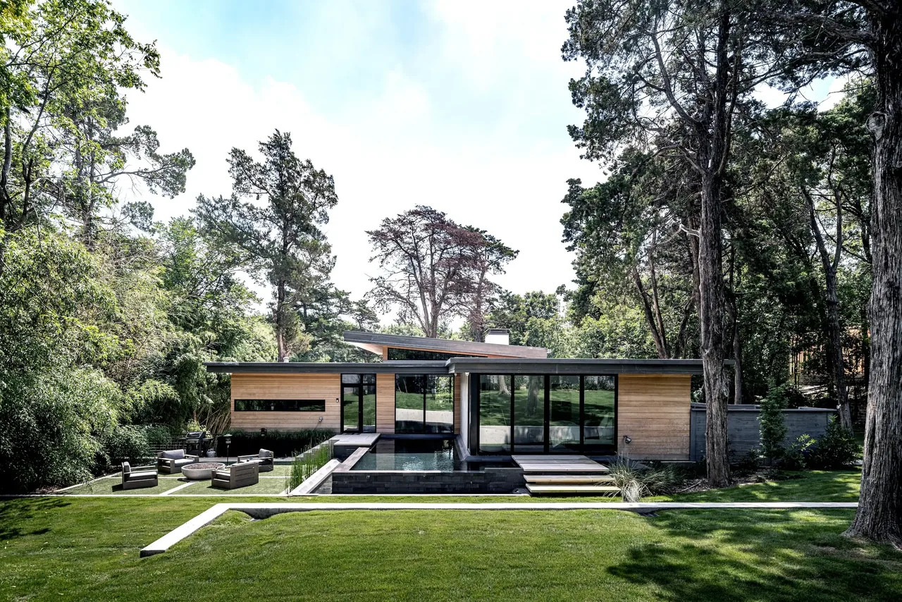 Exterior rear view of a modern bungalow featuring wood siding, floor-to-ceiling glazing, and a reflecting pool in a wooded setting.