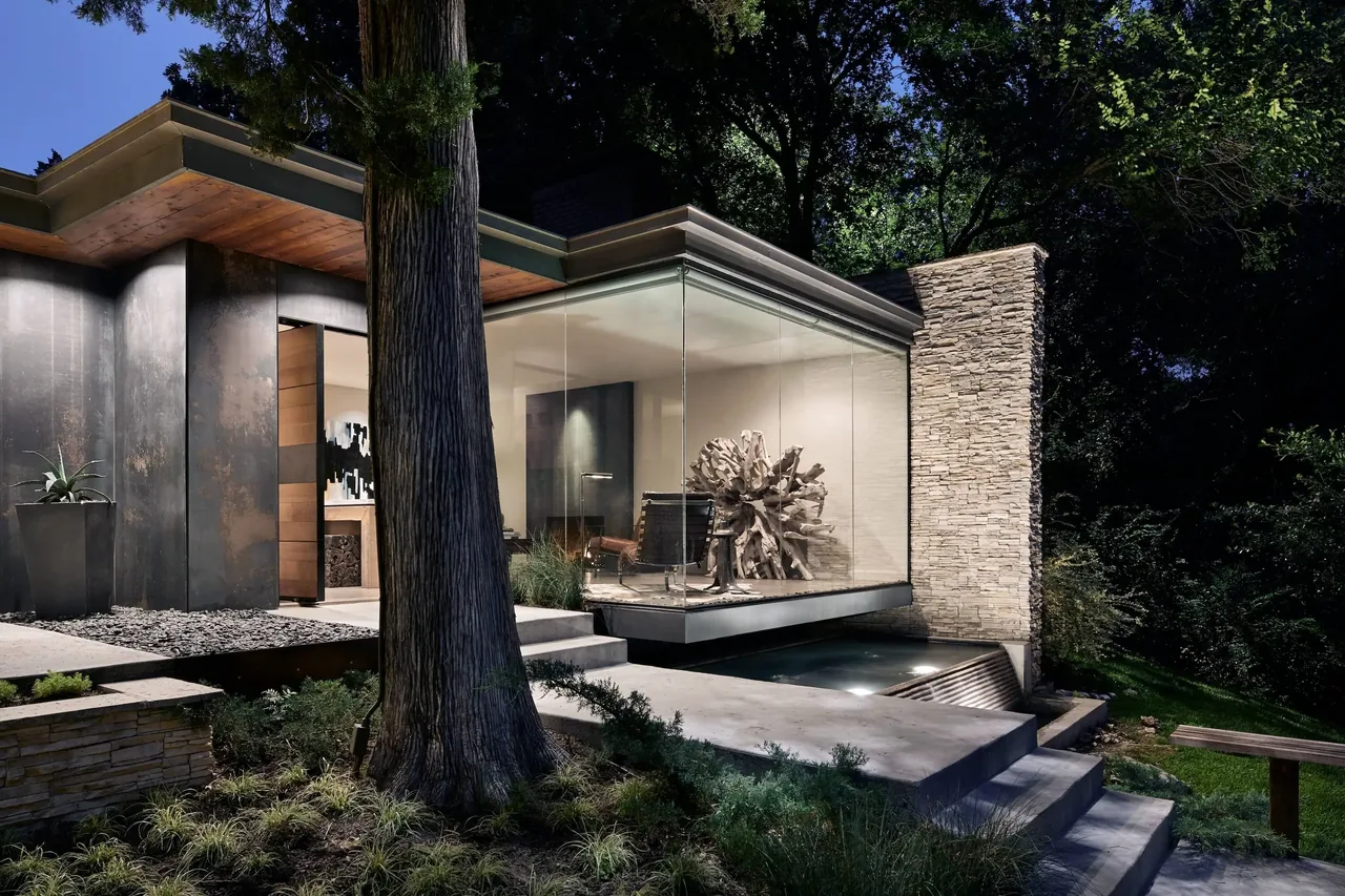 Modern home's corner featuring floor-to-ceiling glazing and a stone wall, shown at night with landscape lighting.