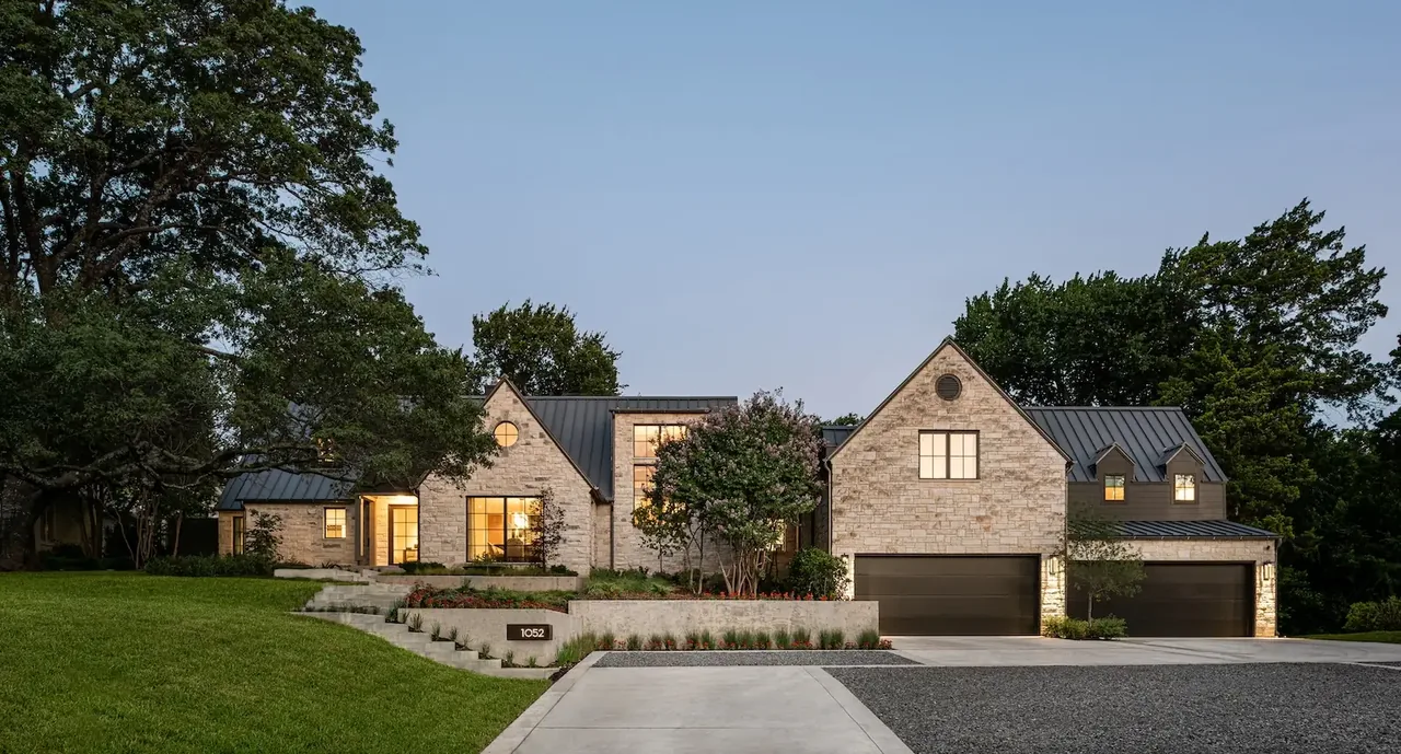 Exterior front view of a modern farmhouse-style house with stone siding and metal gable roofs at dusk.
