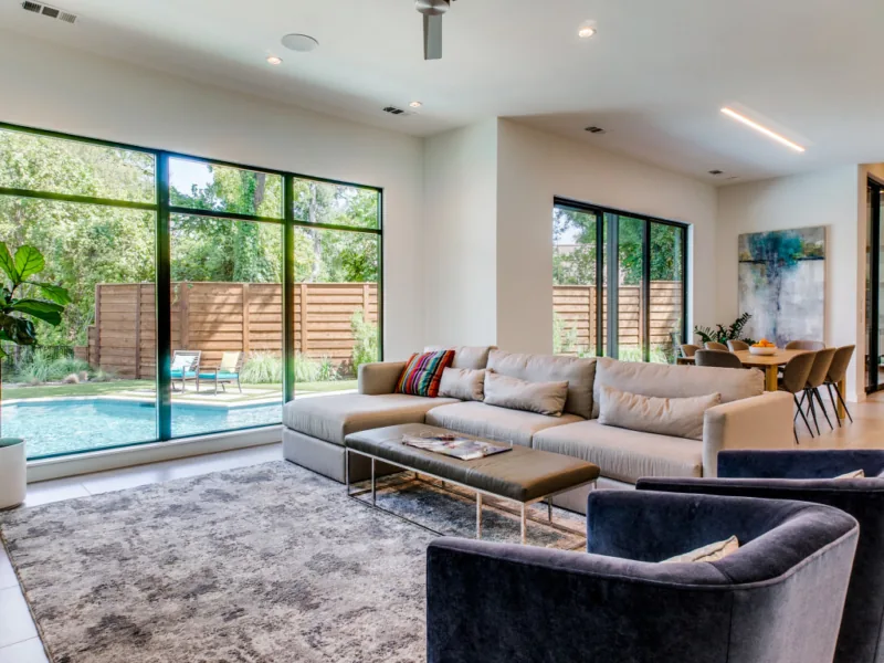 Living area with sectional sofa and black-framed windows showing a fenced yard and pool in the Riverview project.