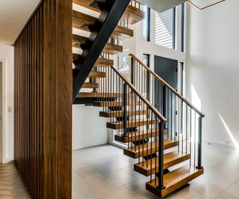 Open riser wood stair with black metal balusters and handrail beside a vertical wood slat wall at entry.