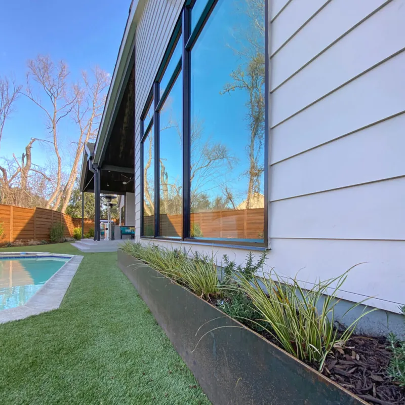 Riverview project exterior shows black window frames, raised planter, artificial grass, and poolside area with wood fence.