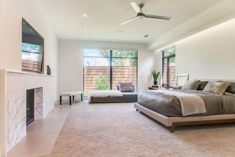 Modern bedroom features a low platform bed, marble fireplace surround, and large black-framed windows overlooking a fenced yard.