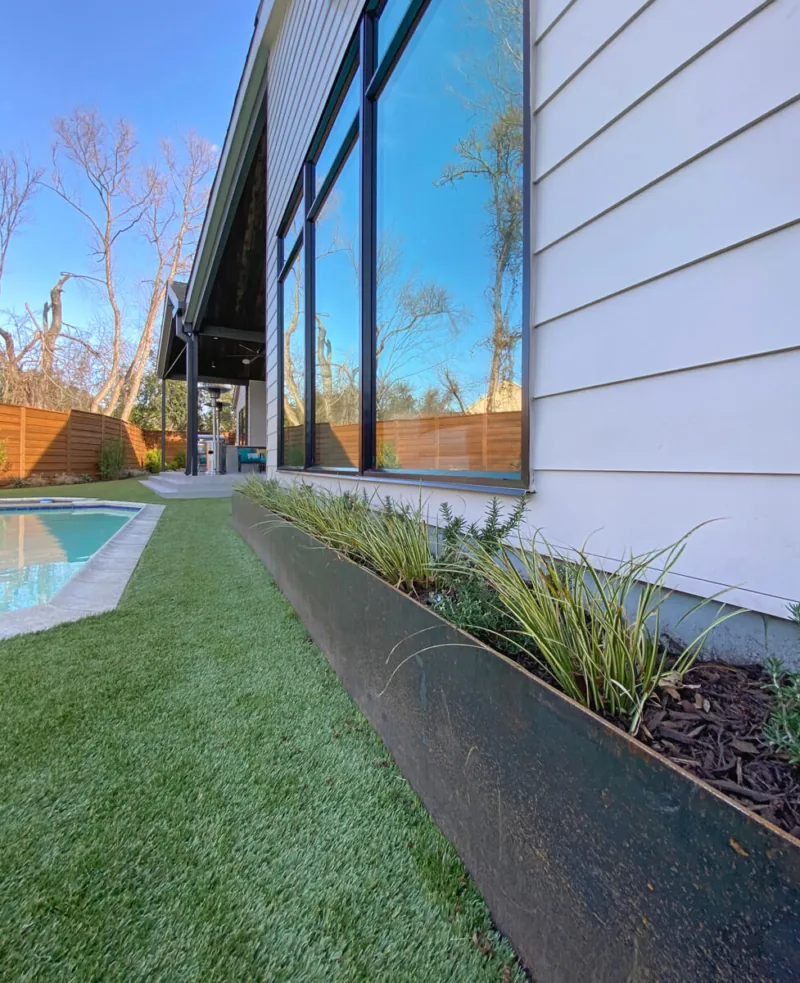 Modern house exterior with large black window frames, metal planter, artificial turf, and pool area.