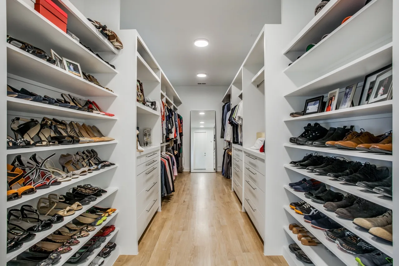 This walk-in closet features white built-in shelving and drawers, with wood flooring and recessed ceiling lights.