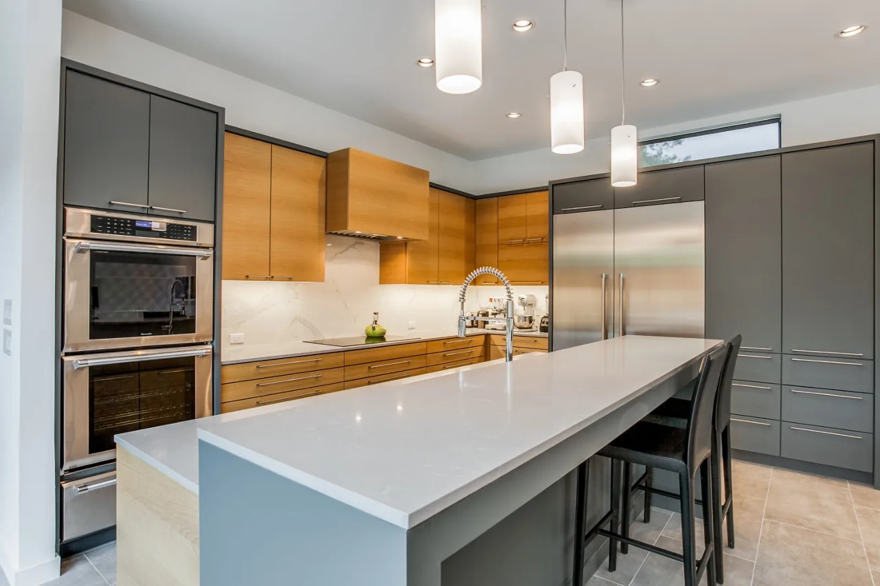A kitchen featuring a large white island, wooden and gray cabinets, and stainless steel built-in appliances under recessed and pendant lighting.
