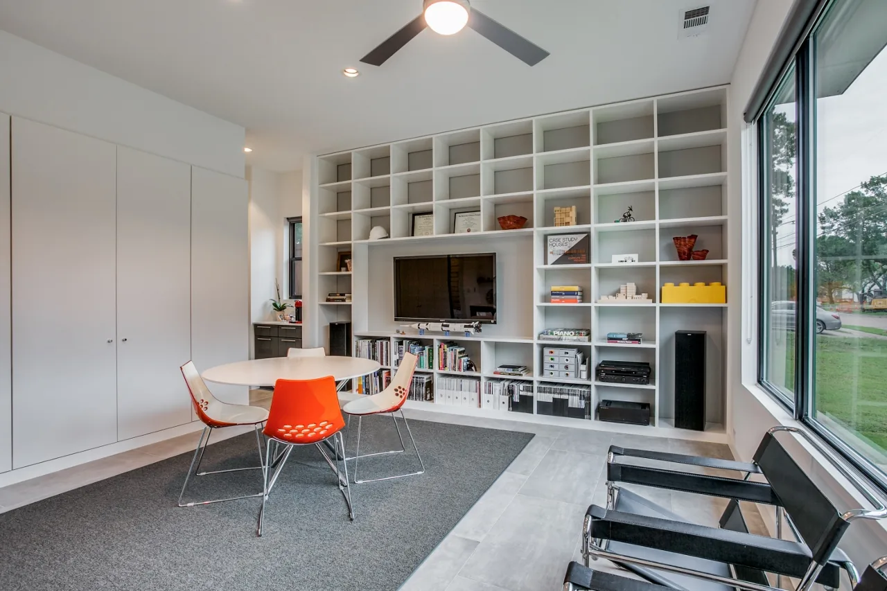 Office space featuring a large white built-in shelving unit with a mounted TV and a round table with red and white chairs on a gray carpet.